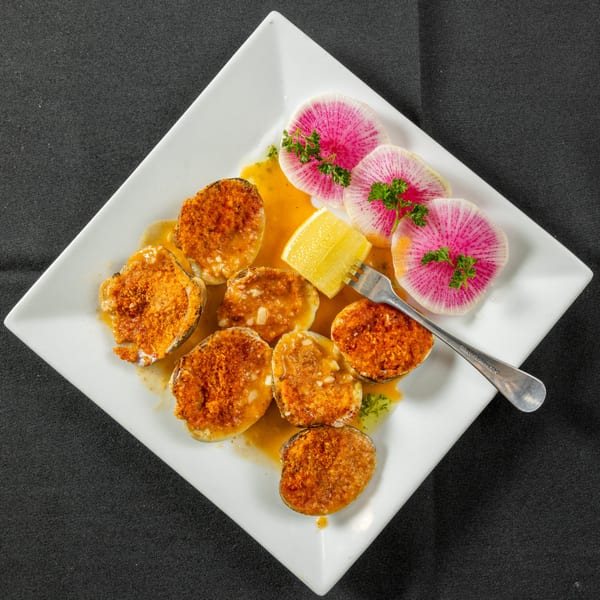 Baked clams with breadcrumbs, lemon, and sliced radishes on a white square plate.