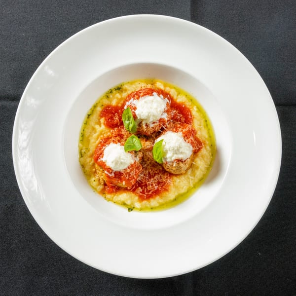 Three meatballs with tomato sauce, mozzarella, and basil on a bed of polenta in a white bowl.