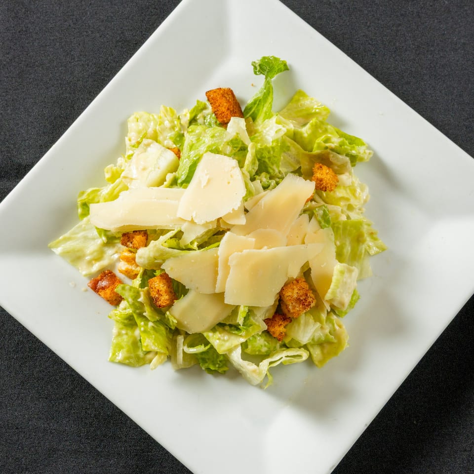 Caesar salad with romaine lettuce, croutons, and shaved Parmesan cheese on a white square plate.