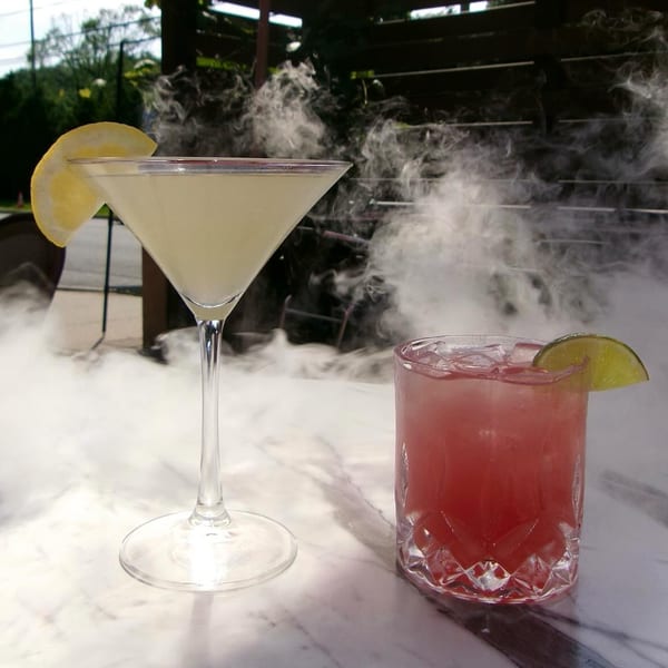 Two cocktails, one yellow martini with lemon and one red drink with lime, surrounded by mist.